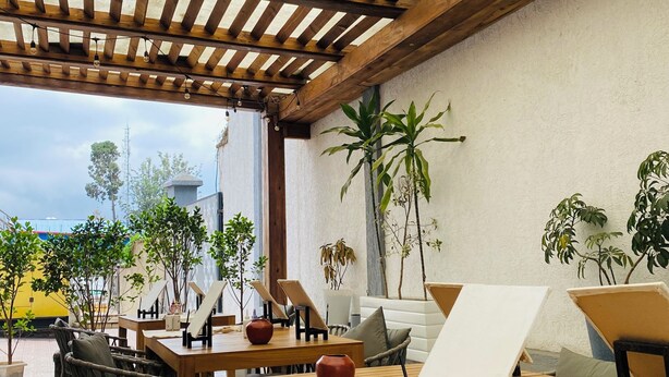 Un espace extérieur aménagé en terrasse, avec des tables et des chaises en bois, des plantes en pot et une pergola illuminée de guirlandes lumineuses.