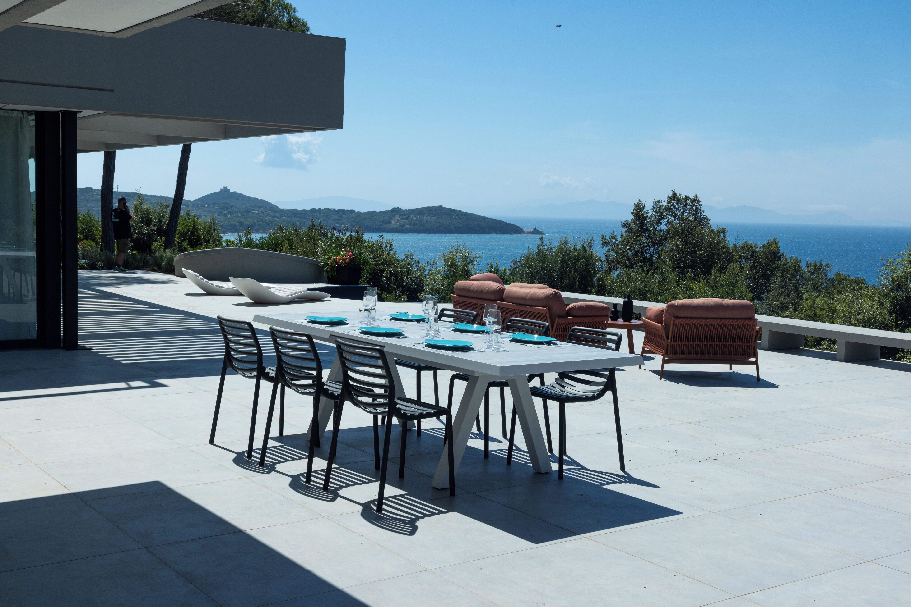 An outdoor dining area with a table set for four, surrounded by chairs and cushions, overlooking a scenic view of the sea and distant hills.