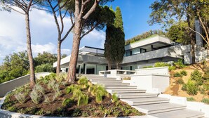 Una casa moderna con una escalera que conduce a una terraza plana, rodeada de jardines ajardinados y árboles.
