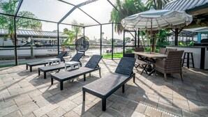 An outdoor patio with lounge chairs, a table, and a grill area, surrounded by a metal fence and palm trees. - Resort-Style Canal Home w/ Pool & Putting Green - Baywatch (Apollo Beach)