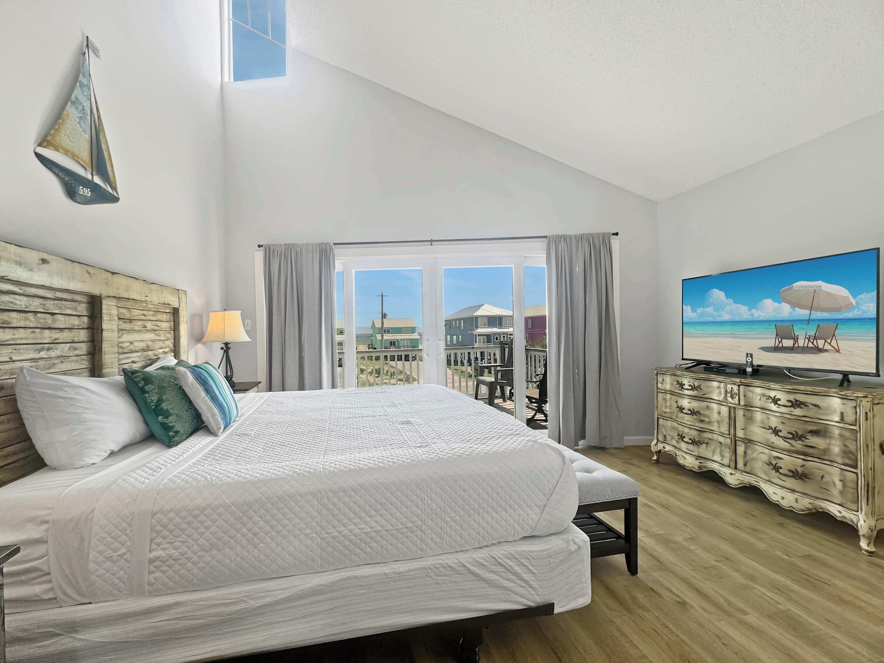 Ein Schlafzimmer mit einem großen Bett, einer Kommode, einem Fernseher, der eine Strandansicht zeigt, und Blick auf einen Balkon mit Geländer.