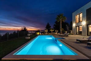 A rectangular swimming pool with a view of the city at dusk, a modern house with a balcony, and a palm tree. - Welcome to Villa El Capitano – Your Private Seaview Retreat Near Split (Podstrana)