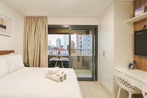 Une chambre d’hôtel équipée d’un lit, d’un bureau, d’une chaise, d’une télévision et d’un balcon donnant sur des immeubles.