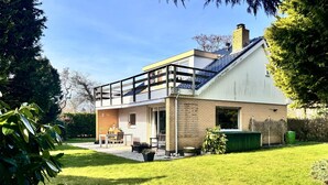 Ein modernes, zweistöckiges Haus mit Balkon, einer Terrasse mit Gartenmöbeln und einem gepflegten Rasen.