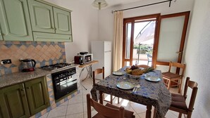 A kitchen with green cabinets, a stove, and a microwave on a countertop. There is a dining table with chairs and a fruit bowl. - La Casetta Di Greise (Budoni)