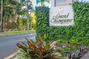 A sign reading "Grand Champions" with a lush green hedge backdrop and tropical plants in the foreground.