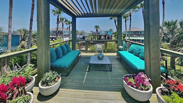 Una terraza de madera con zona de estar cubierta, rodeada de macetas con plantas y vistas a una zona residencial.