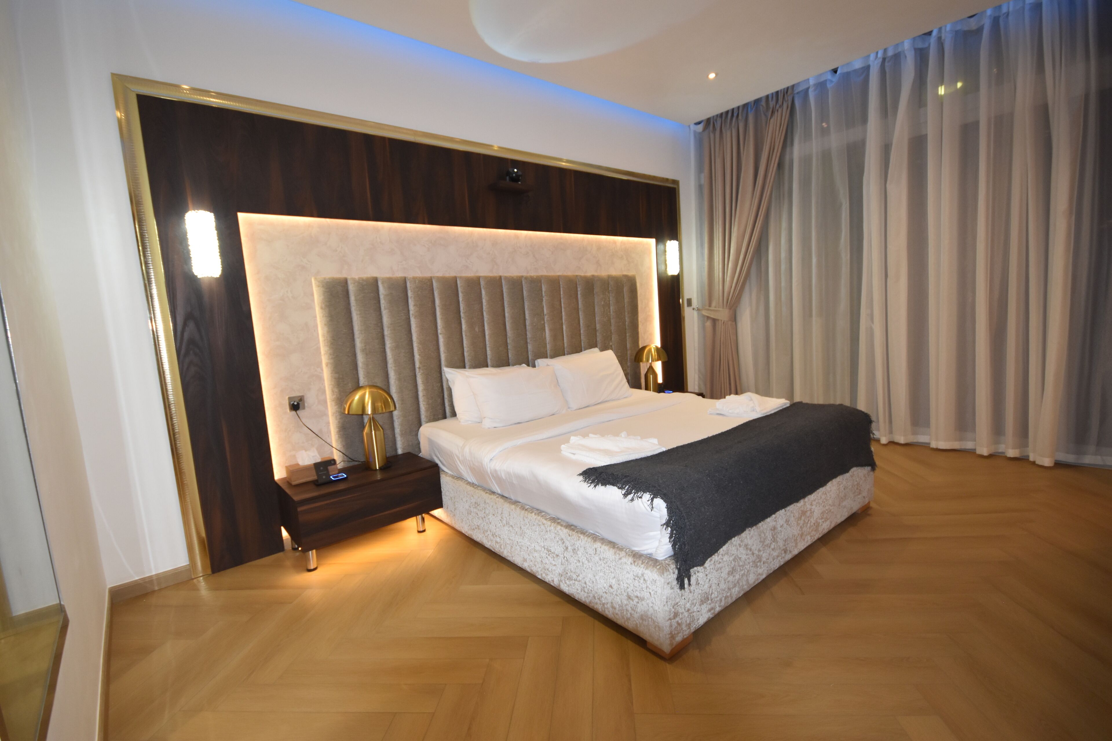 A modern hotel room with a large bed, wooden flooring, and a headboard with built-in lighting.