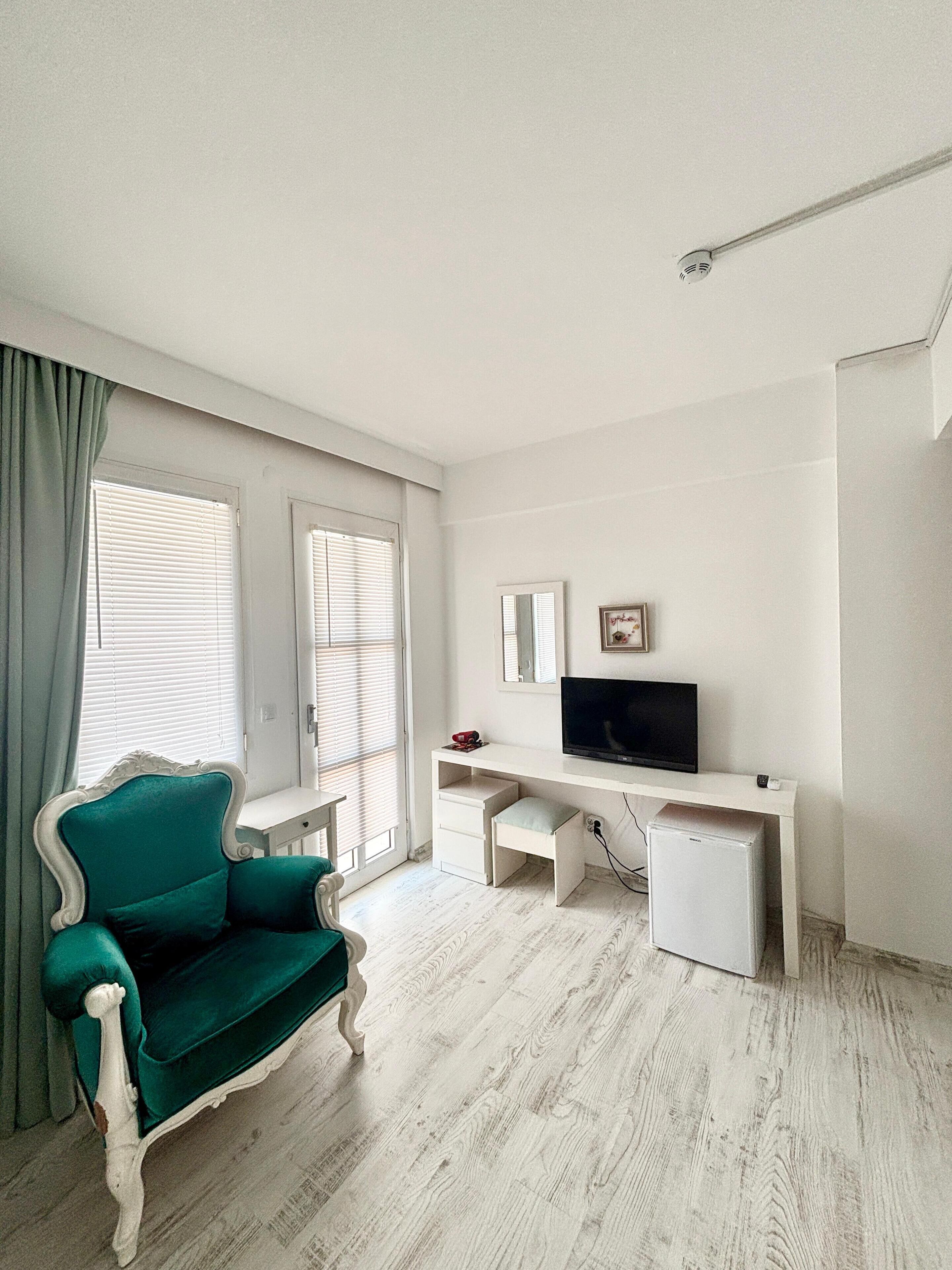 Une pièce avec un fauteuil turquoise, un bureau blanc avec un téléviseur et un petit réfrigérateur blanc.