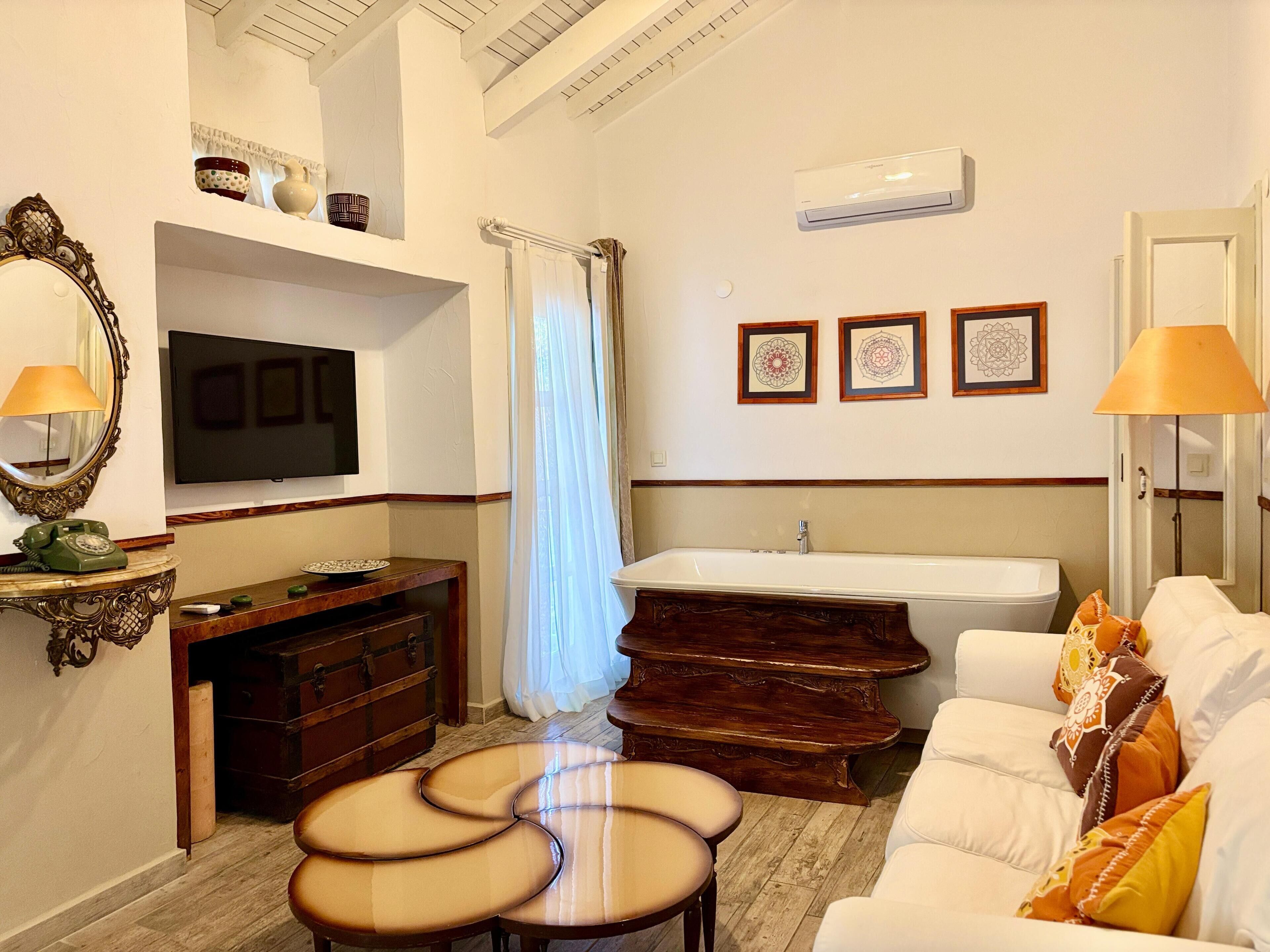 A hotel room with a TV, a wooden dresser, a vanity with a mirror, a bathtub, a white sofa, and a dining table with chairs.