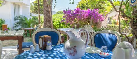 An outdoor dining area with a blue-striped tablecloth, white ceramic tableware, and a view of a white building and purple flowers in the background.