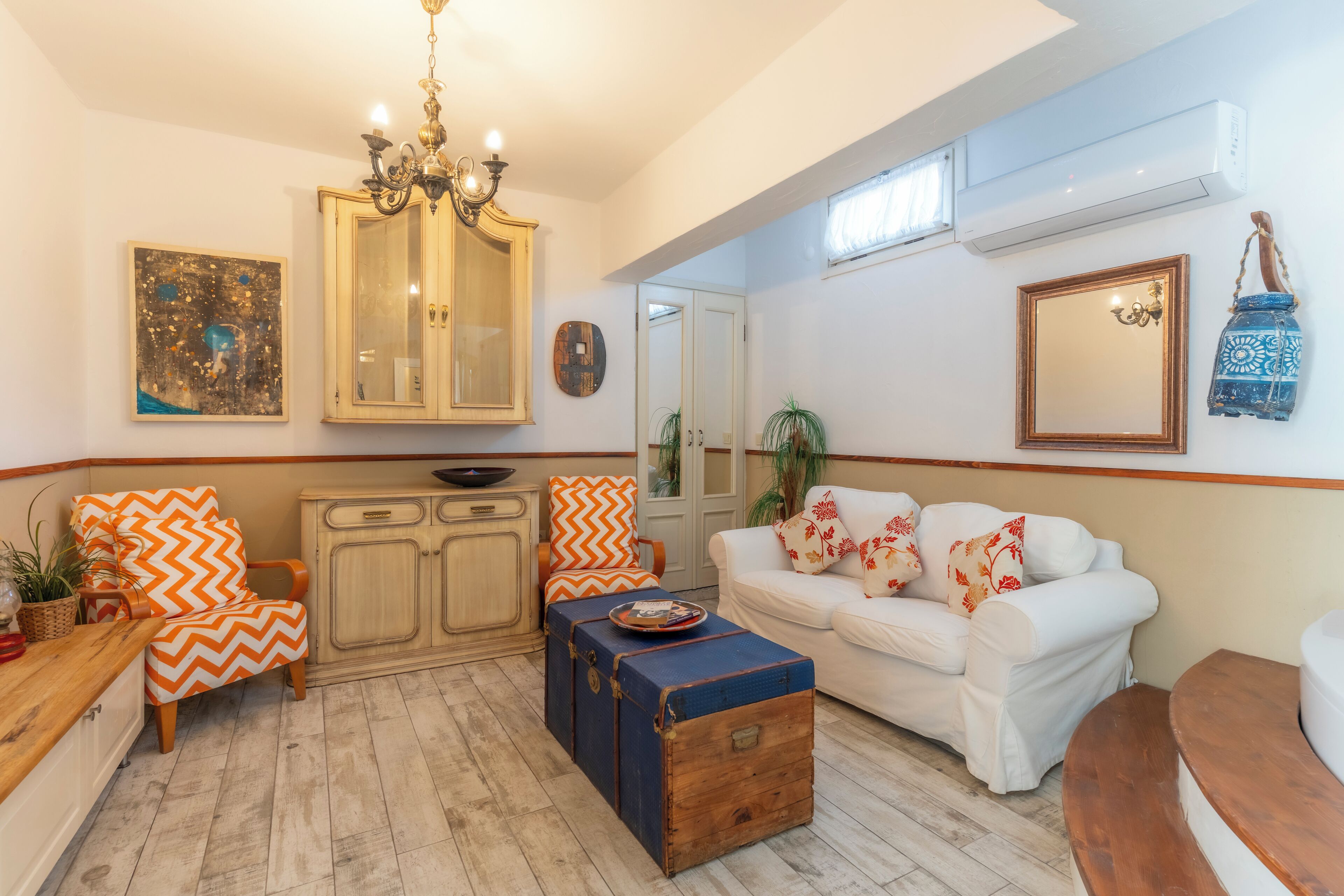 A living room with a white sofa, a blue trunk, a wooden cabinet, and a chandelier.
