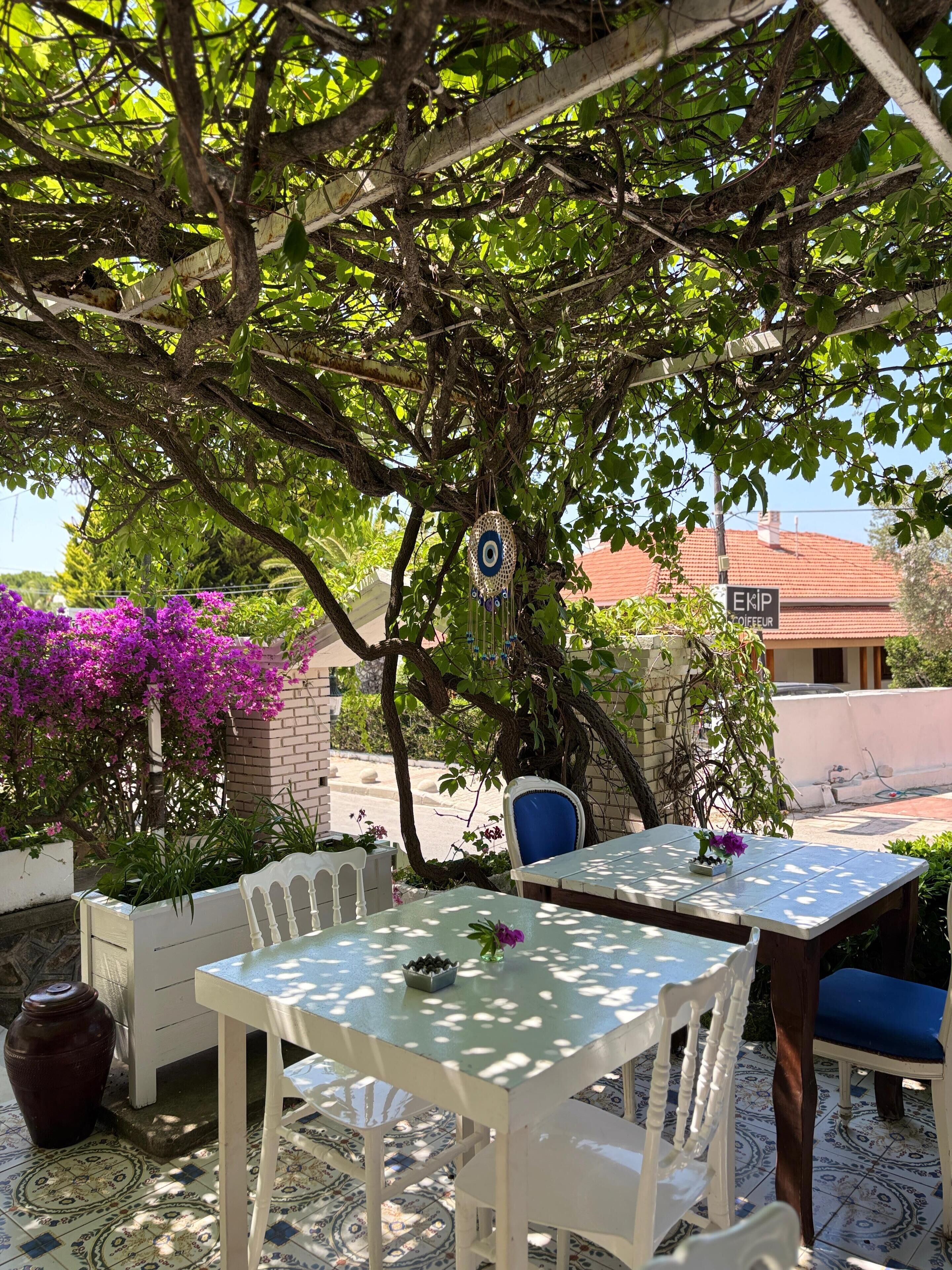 An outdoor seating area with white tables and chairs, surrounded by lush greenery and purple bougainvillea flowers.