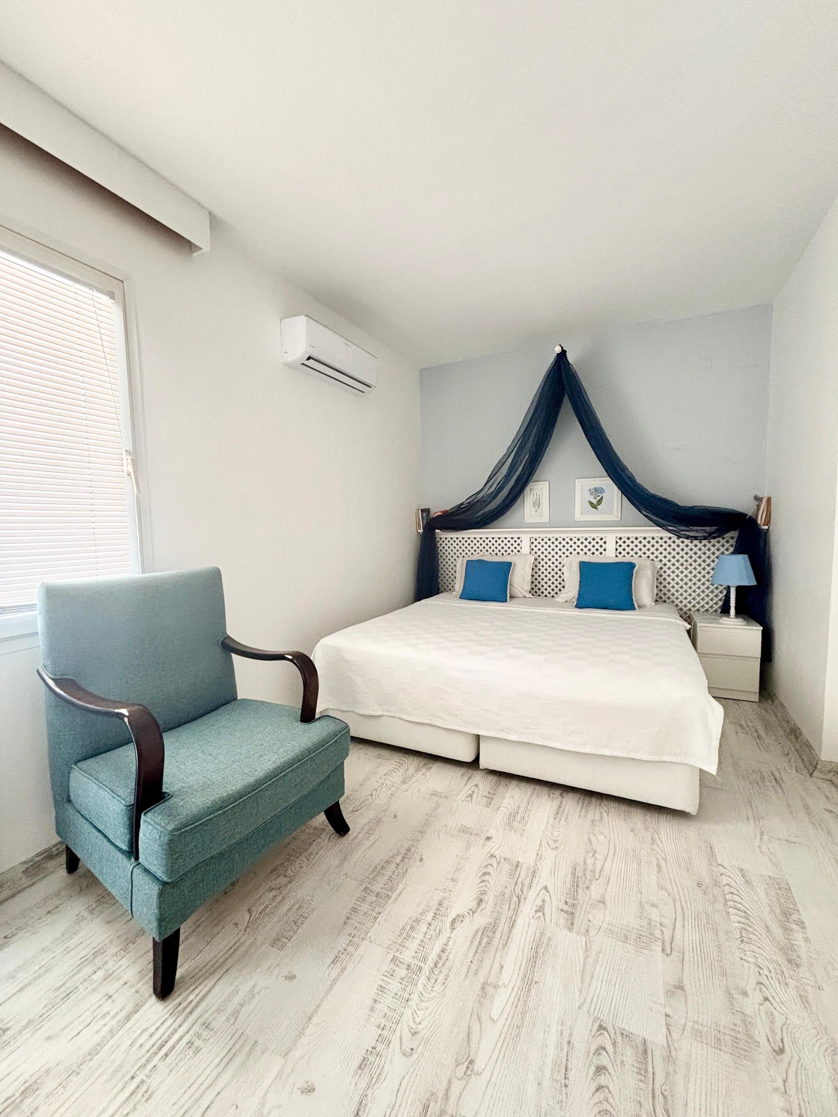 A bedroom with a bed, a chair, a nightstand, and a window with blinds.