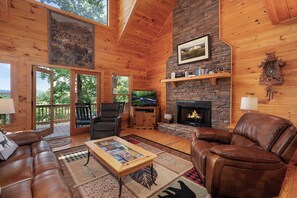 A cozy cabin interior with a fireplace, leather furniture, and a large window overlooking a forest.