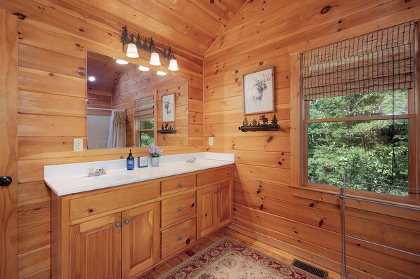 A wooden cabin bathroom with a large mirror, wooden cabinets, a white sink, and a window with a view of greenery.