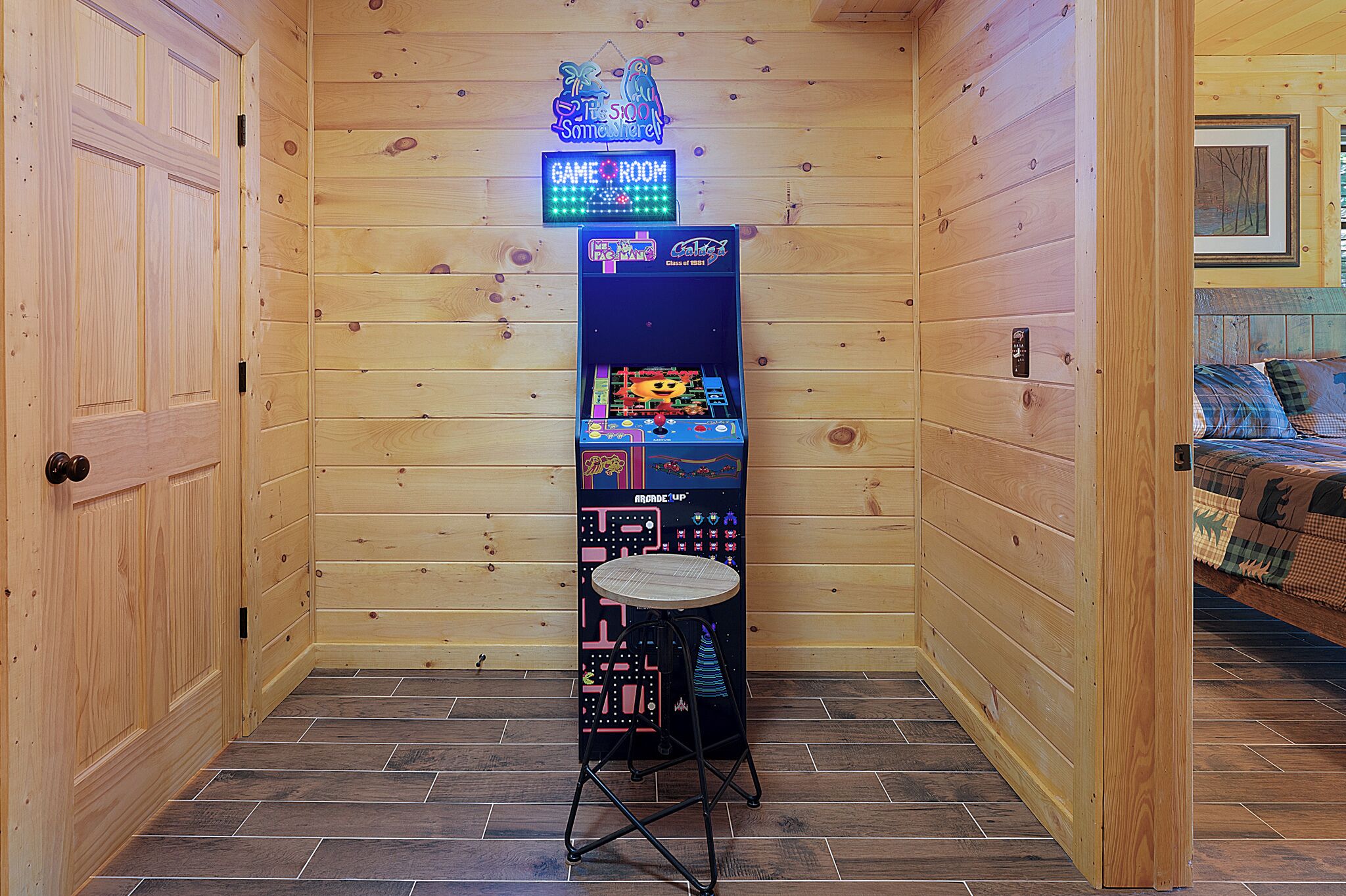 A wooden-paneled room with a neon-lit game room sign, a bed with a plaid blanket, and a small table with a neon sign.