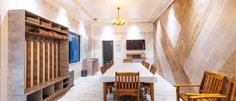 A spacious dining area with a long table and wooden chairs, a built-in bench with storage, a television, and a kitchen area in the background.