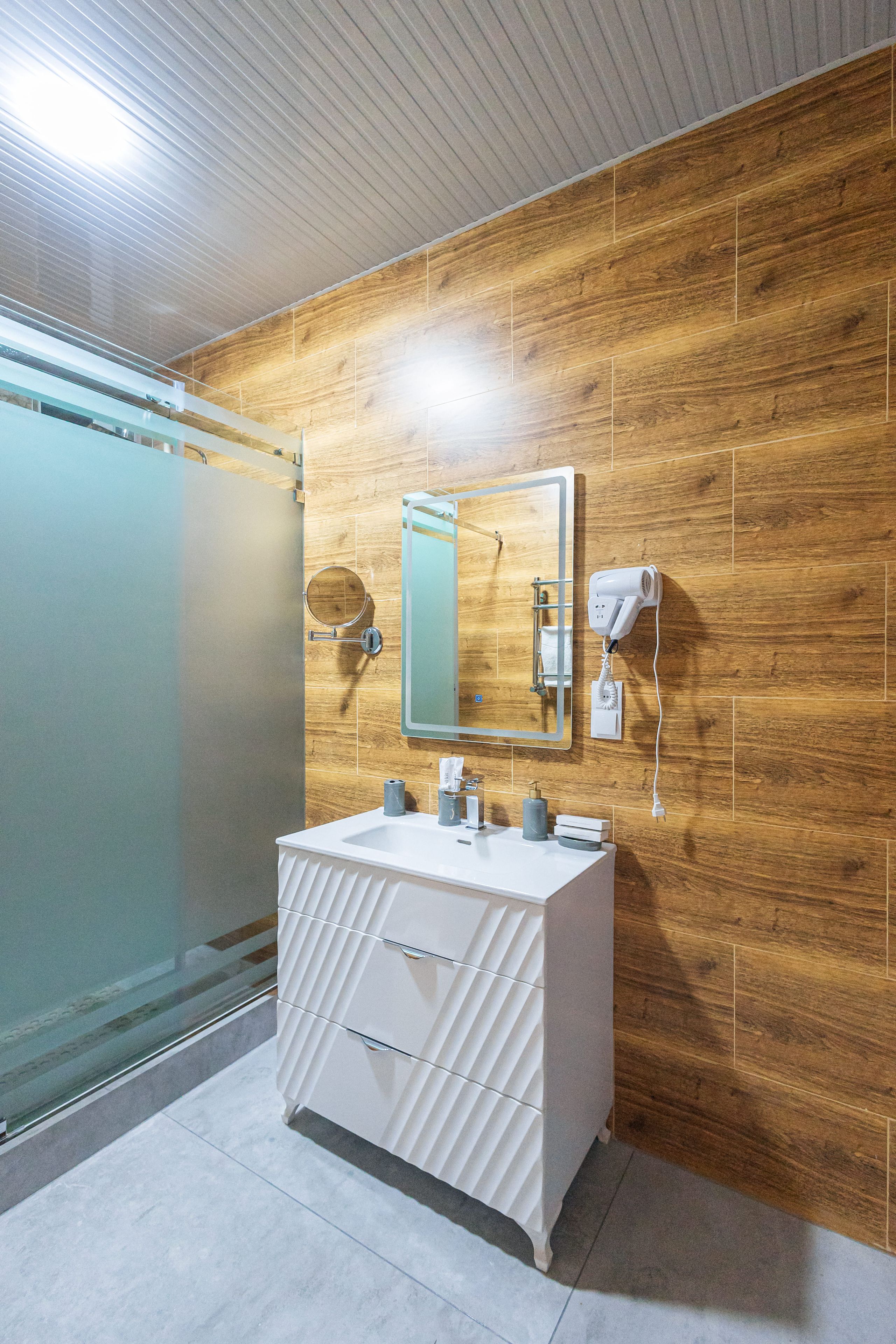 Un bany modern amb una paret de fusta, un lavabo blanc, un mirall, un assecador d'eixugat i una cabina de dutxa de vidre.