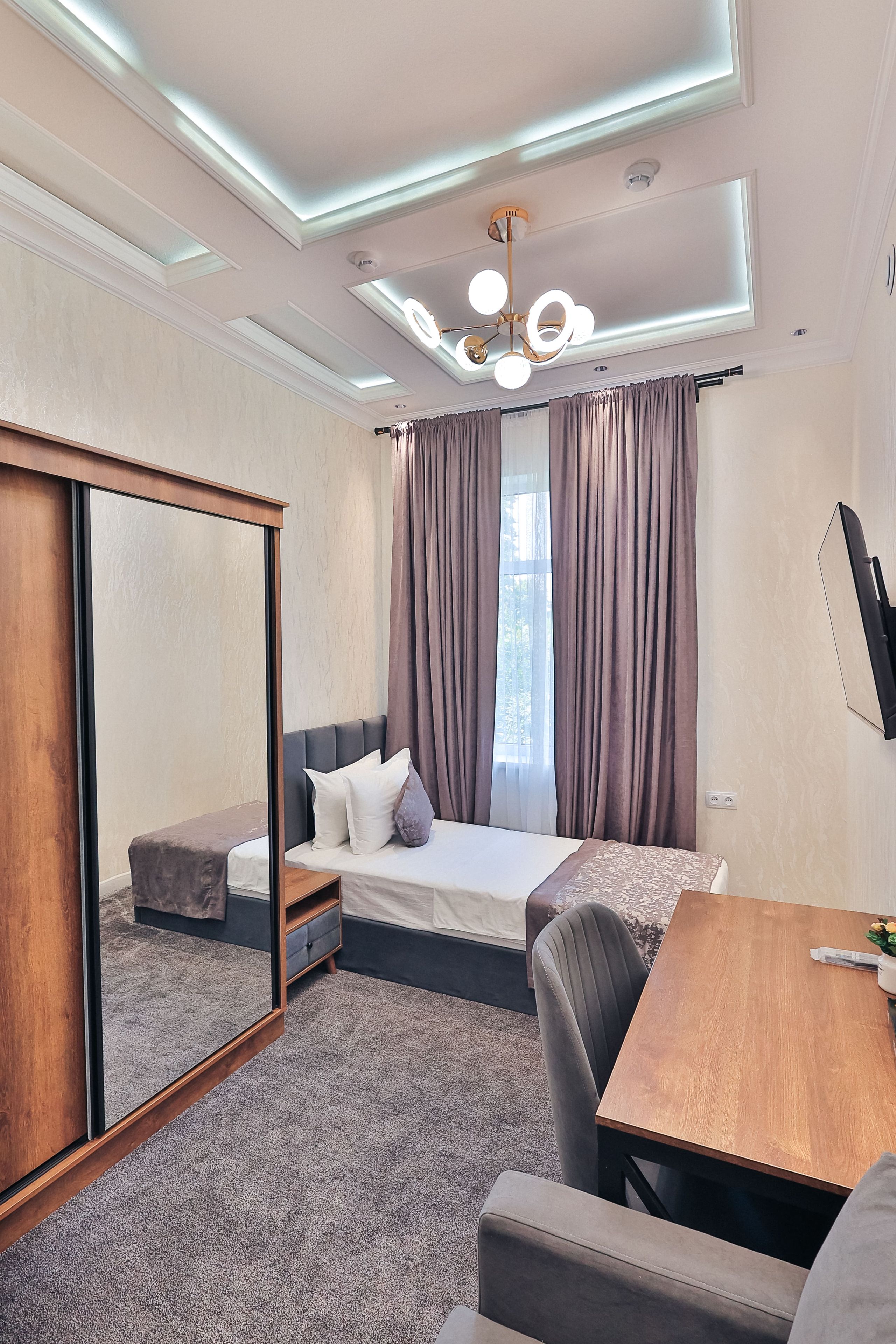 A modern hotel room with a bed, a desk, a chair, a mirror, and a television.