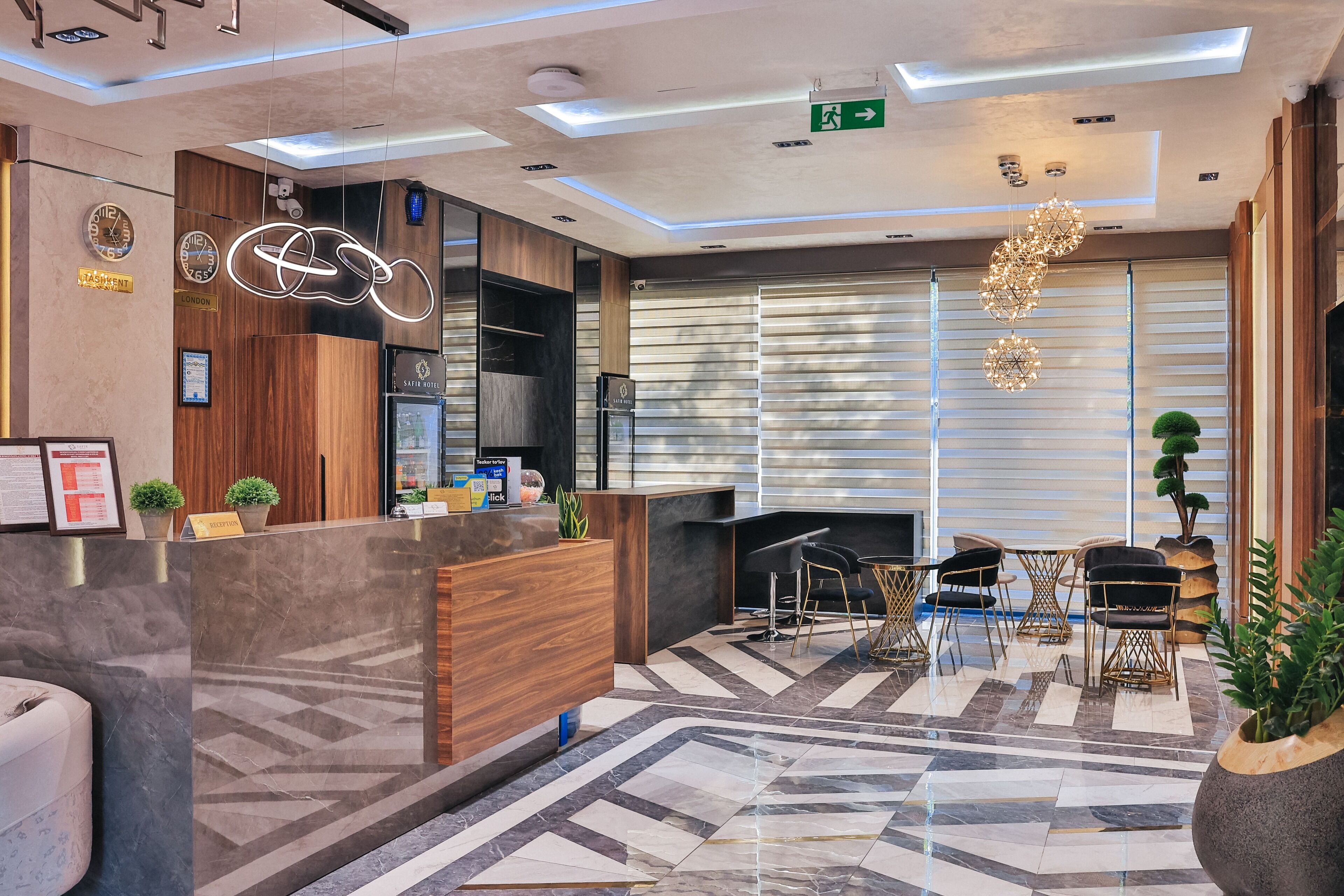 A modern reception area with a wooden reception desk, a digital clock, and a reception sign. There are potted plants, a seating area with chairs, and a patterned floor.