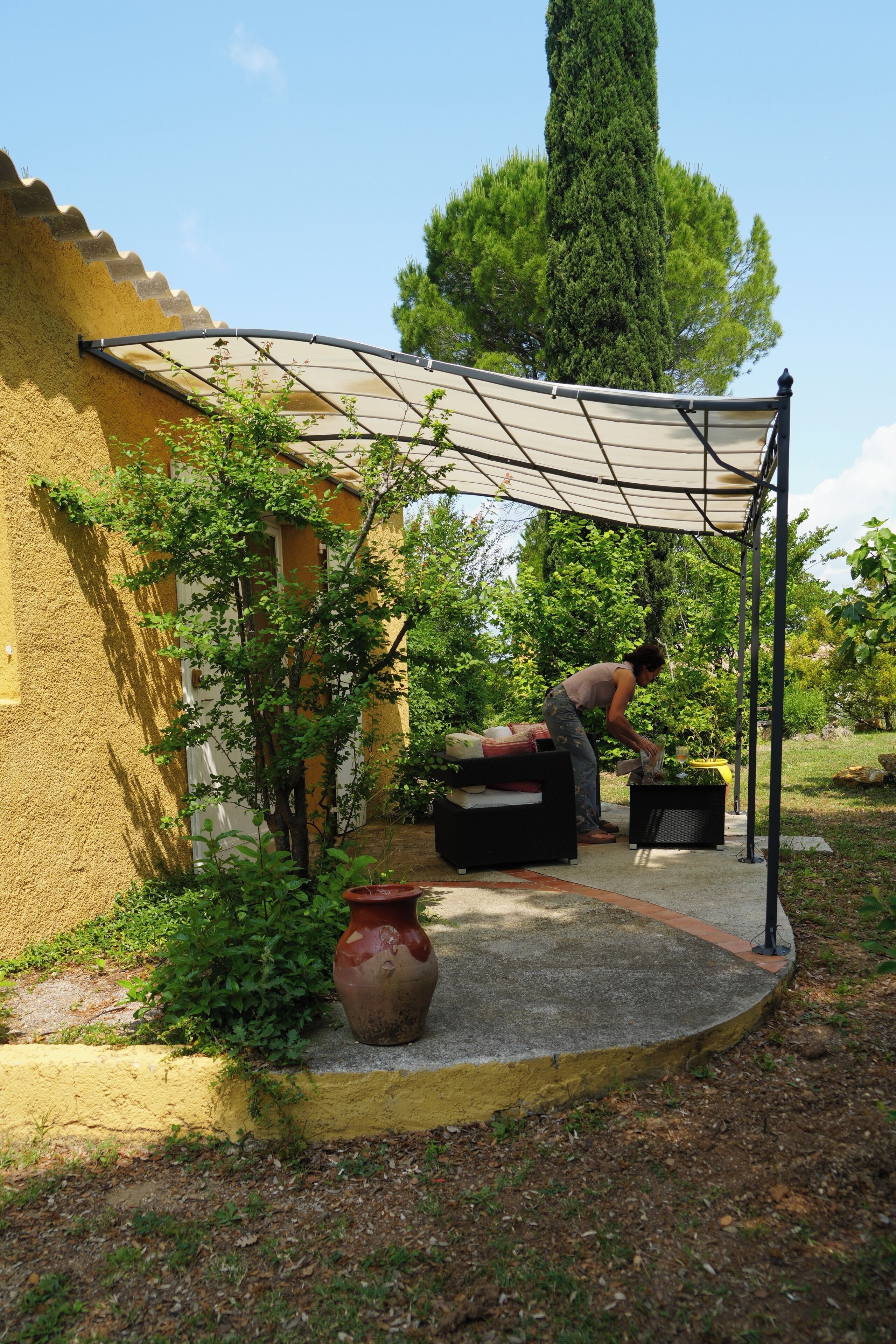 A covered outdoor area with a yellow building, a large tree, and a person tending to plants.