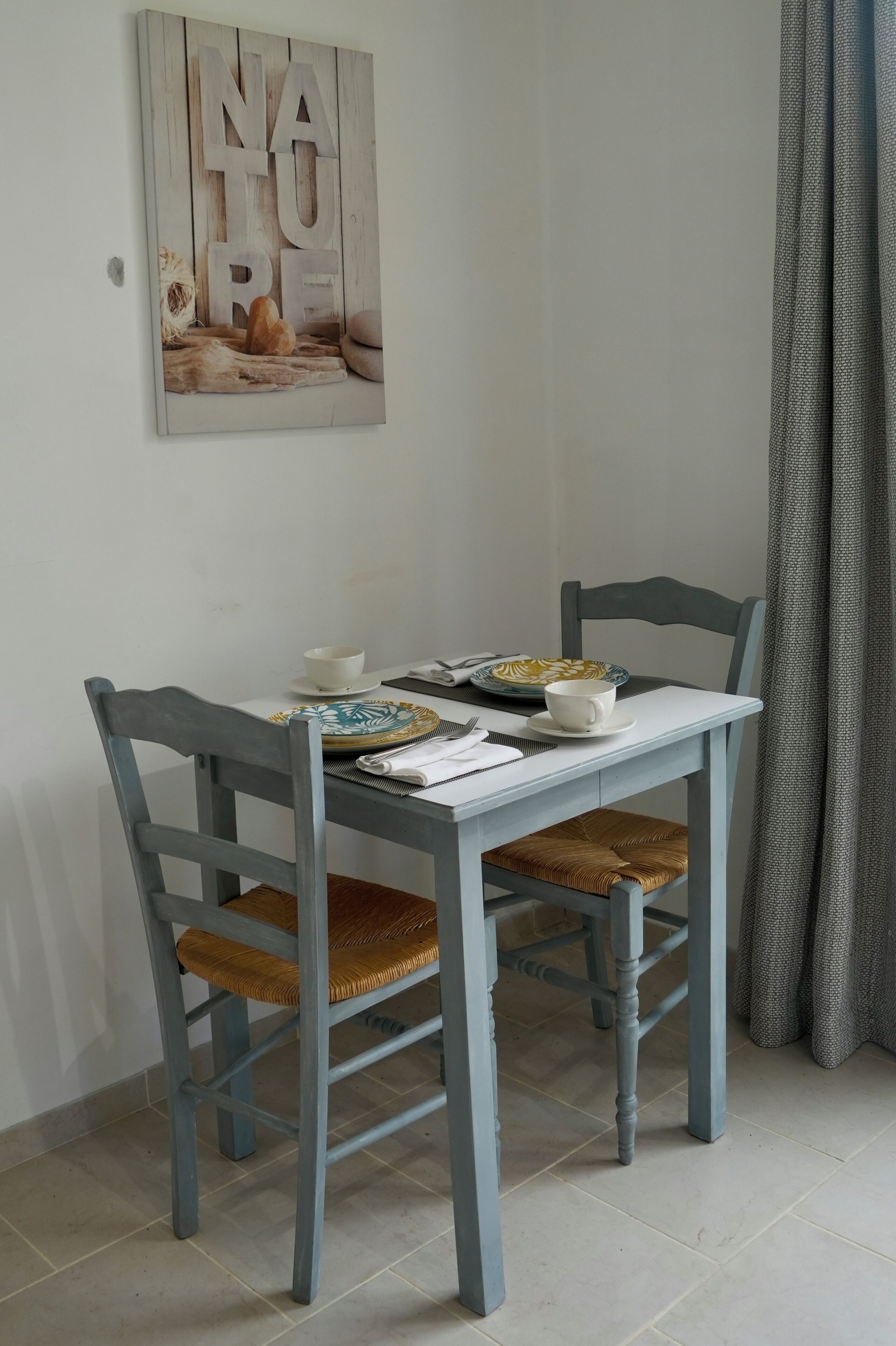 Un coin repas avec une table dressée pour deux personnes, agrémentée d’une nappe blanche, de deux assiettes, d’un bol et d’une tasse. Les chaises sont recouvertes d’un tissu clair et dotées de coussins d’assise tissés. Une œuvre d’art rustique représentant le mot « NATURE » est visible au-dessus de la table.