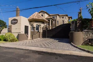 Un grand immeuble à plusieurs étages, doté d’une entrée fermée par un portail, d’une petite annexe avec un toit en tuiles et d’un mur de pierre agrémenté d’un jardin.