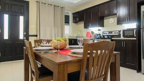 A dining area with a wooden table, chairs, and a fruit bowl. There is a kitchen in the background with dark cabinets and appliances. - Star Tree- 2 Bedroom Condo 10 mins to New Kingston (Kingston)