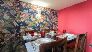 Mesa de jantar posta com pratos, copos e talheres em um quarto com papel de parede floral e uma parede com detalhes vermelhos.