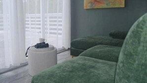 Ein Zimmer mit einem großen Fenster, einer grünen Sofagarnitur, einem weißen Hocker und einem farbenfrohen, abstrakten Gemälde an der Wand.