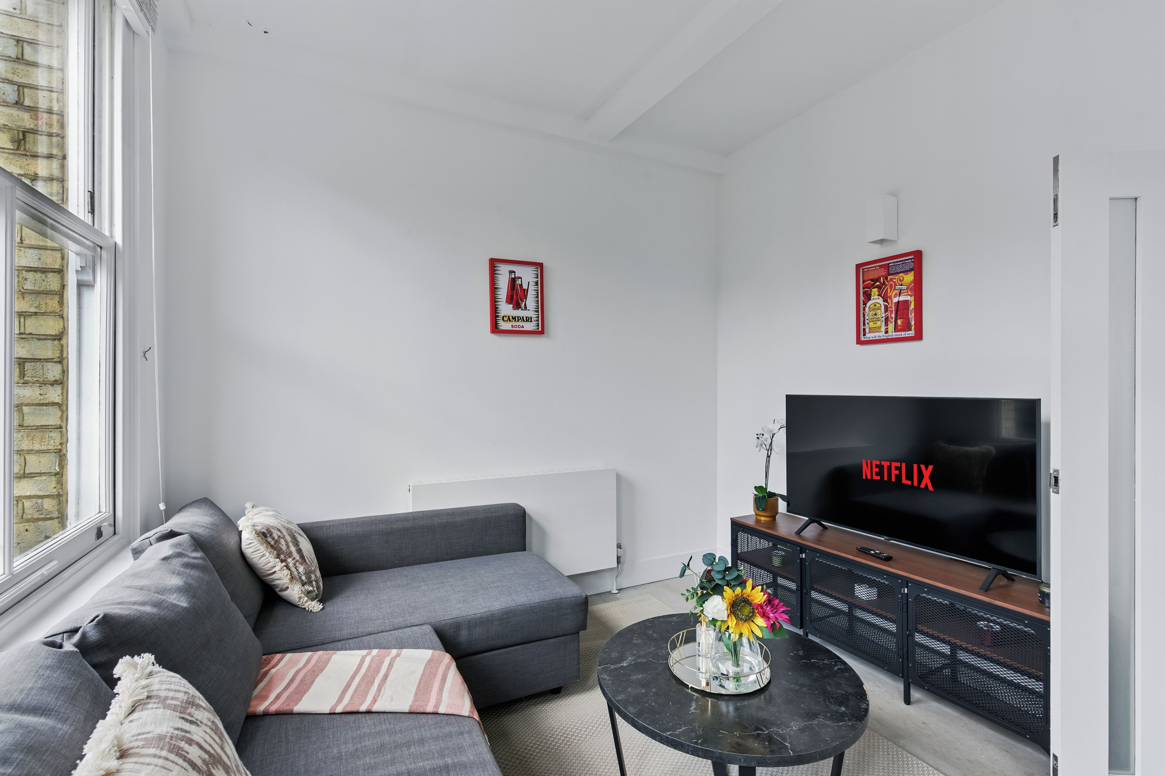 Una sala de estar moderna con un sofá gris, una mesa redonda de centro con un florero, un televisor de pantalla plana y dos carteles enmarcados en la pared.
