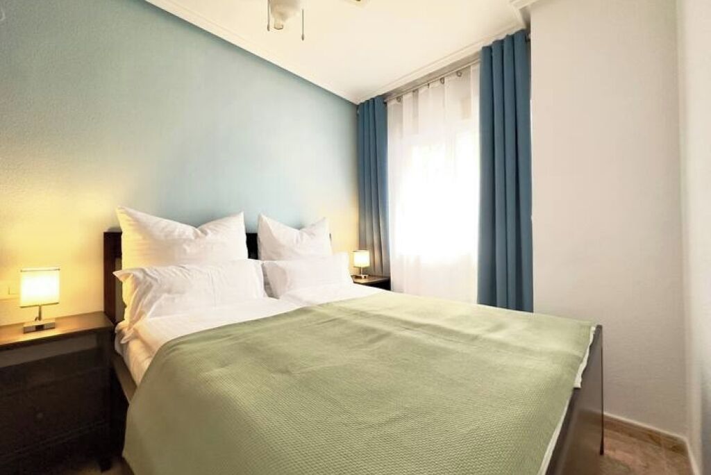 Een netjes opgemaakt bed met witte lakens en een groene deken in een kamer met lichtblauwe muren en een raam met blauwe gordijnen.