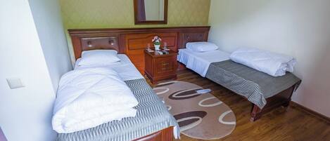 A hotel room with two single beds, a wooden headboard, a small bedside table with a vase of flowers, and a round rug on the wooden floor.
