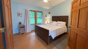 Une chambre à coucher avec un lit en bois, un ventilateur de plafond, une fenêtre avec des rideaux, une table de chevet avec une lampe et une porte.