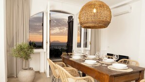 Een eetruimte met een houten tafel voor zes personen, rieten stoelen, een grote geweven hanglamp en uitzicht op de ondergaande zon door een raam.