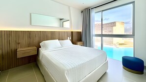 Ein modernes Hotelzimmer mit einem großen Bett, einem hölzernen Kopfteil, einem Nachttisch, einer blauen Bank und einem großen Fenster mit Blick auf Gebäude.