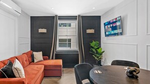 A modern living room with a sofa, a dining table with chairs, a TV mounted on the wall, and a window with curtains.