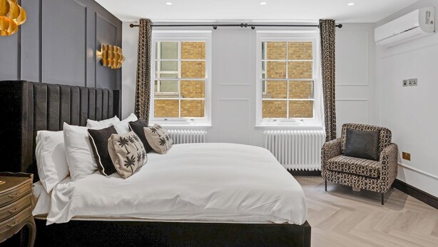 A modern bedroom with a large bed, two windows with curtains, a patterned armchair, and a radiator.