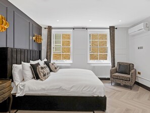 Un dormitorio moderno con una cama grande, dos ventanas con cortinas, un sillón estampado y un radiador.