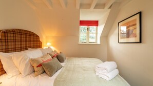 Un dormitorio con cama, almohadas, una ventana con cortinilla roja y un cuadro en la pared.