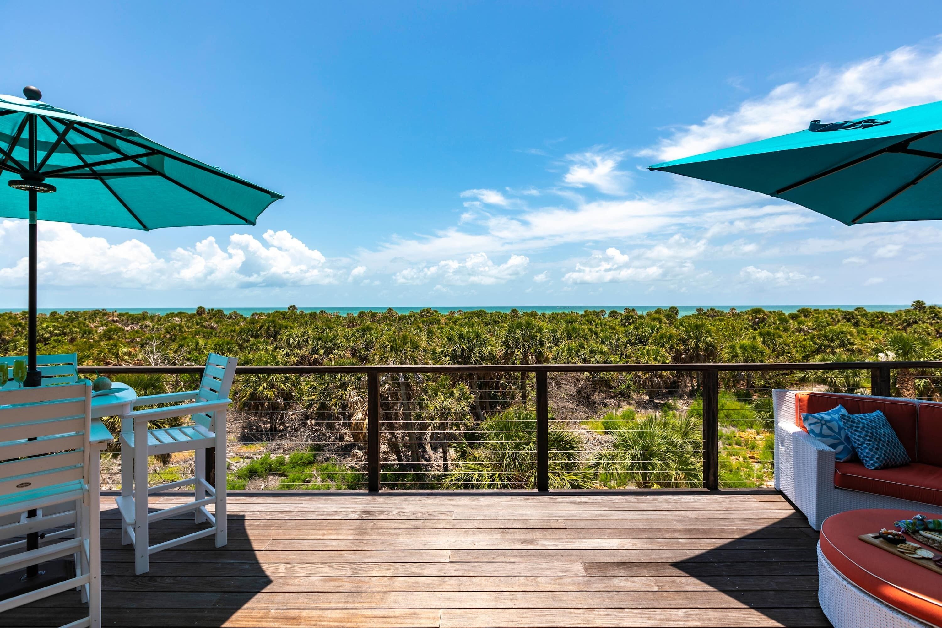 A wooden deck with a white sofa and two turquoise umbrellas, overlooking a lush landscape.