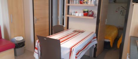Een kleine kamer met een bed, een tafel met een gestreept tafelkleed, een stoel, een kleine prullenbak en een plank met diverse spullen.