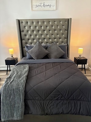 Un dormitorio con cabecera capitonada, dos lámparas de noche, cama con colcha oscura y un cartel en la pared que dice 'sweet dreams'.