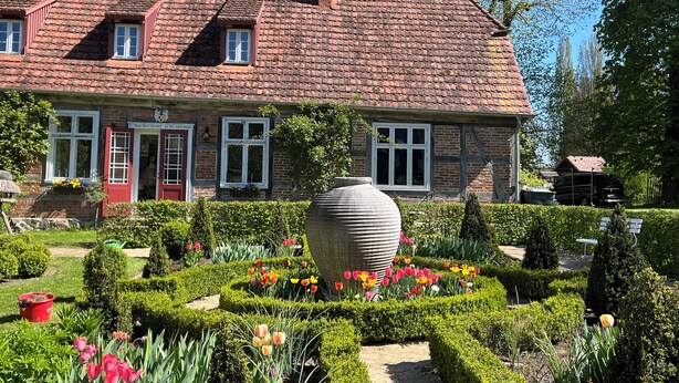 Une maison traditionnelle en briques, avec un toit en tuiles, un grand vase dans le jardin et un parterre de fleurs bien entretenu.