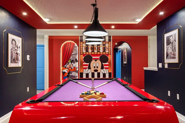 A pool table with a red surface and a cartoon character in the center, surrounded by framed pictures and a 'Tickets' sign. - Mega Themed Villa Near Disney w/ Pool & Arcade (Clermont)