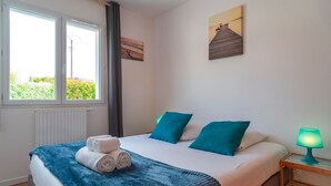 A bedroom with a bed, two pillows, a blanket, a rolled towel, a nightstand with a lamp, and a window with curtains. - La Bulle de Landrezac - Jacuzzi/Proche Plage (Sarzeau)