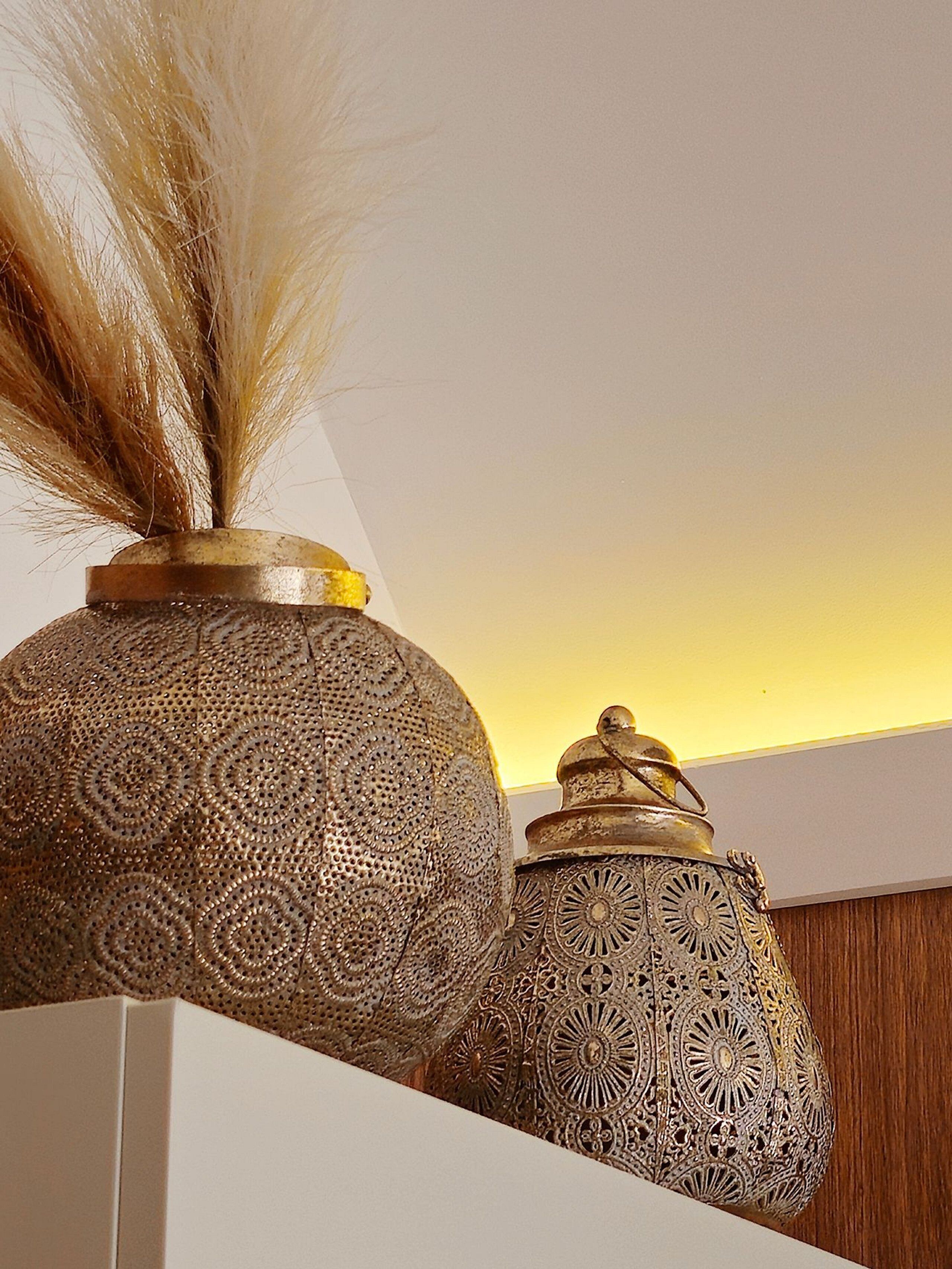 Dos jarrones de metal ornamentados con intrincados diseños, uno con una pluma encima, expuestos sobre un estante blanco frente a un fondo de madera.