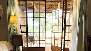 A room with a bed, a small table with a lamp, a glass-top table, and a view through a wooden door to an outdoor area.