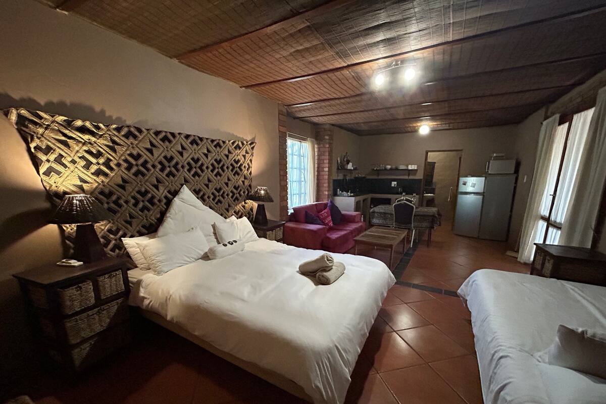 A hotel room with a large bed, a red armchair, a small table, and a kitchenette in the background.